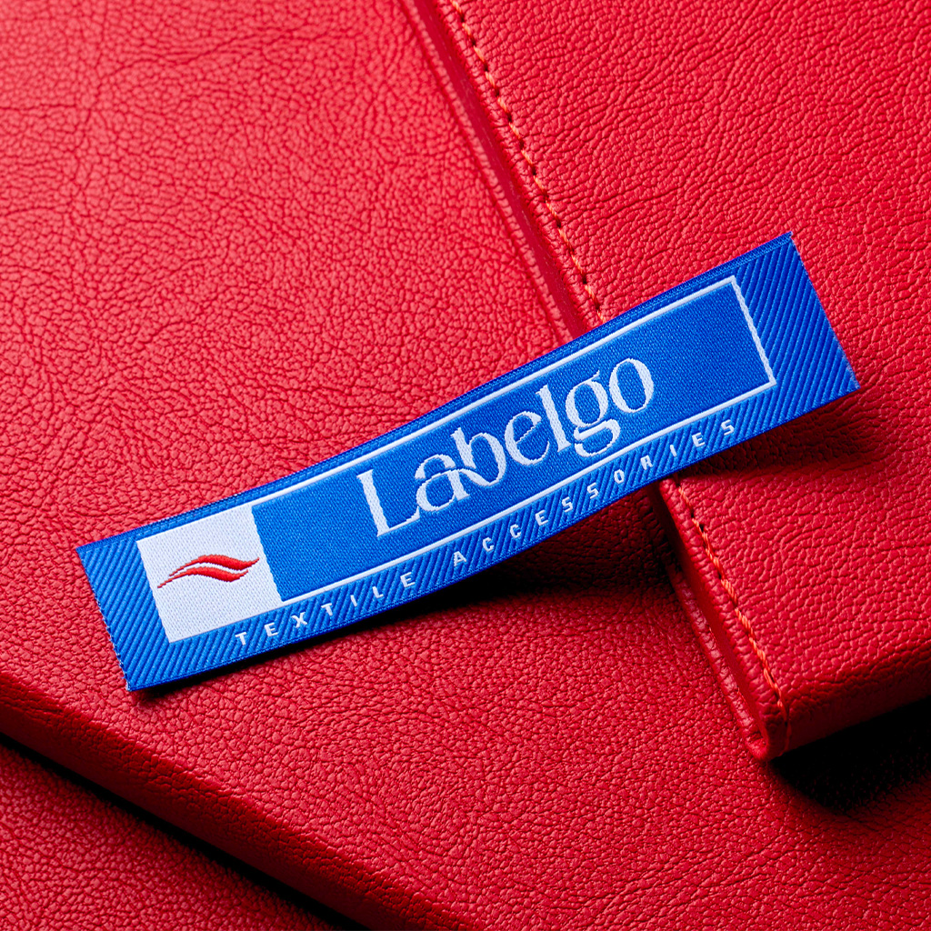 Etiqueta de tela tejida azul con letras blancas fijada sobre una superficie de cuero rojo.