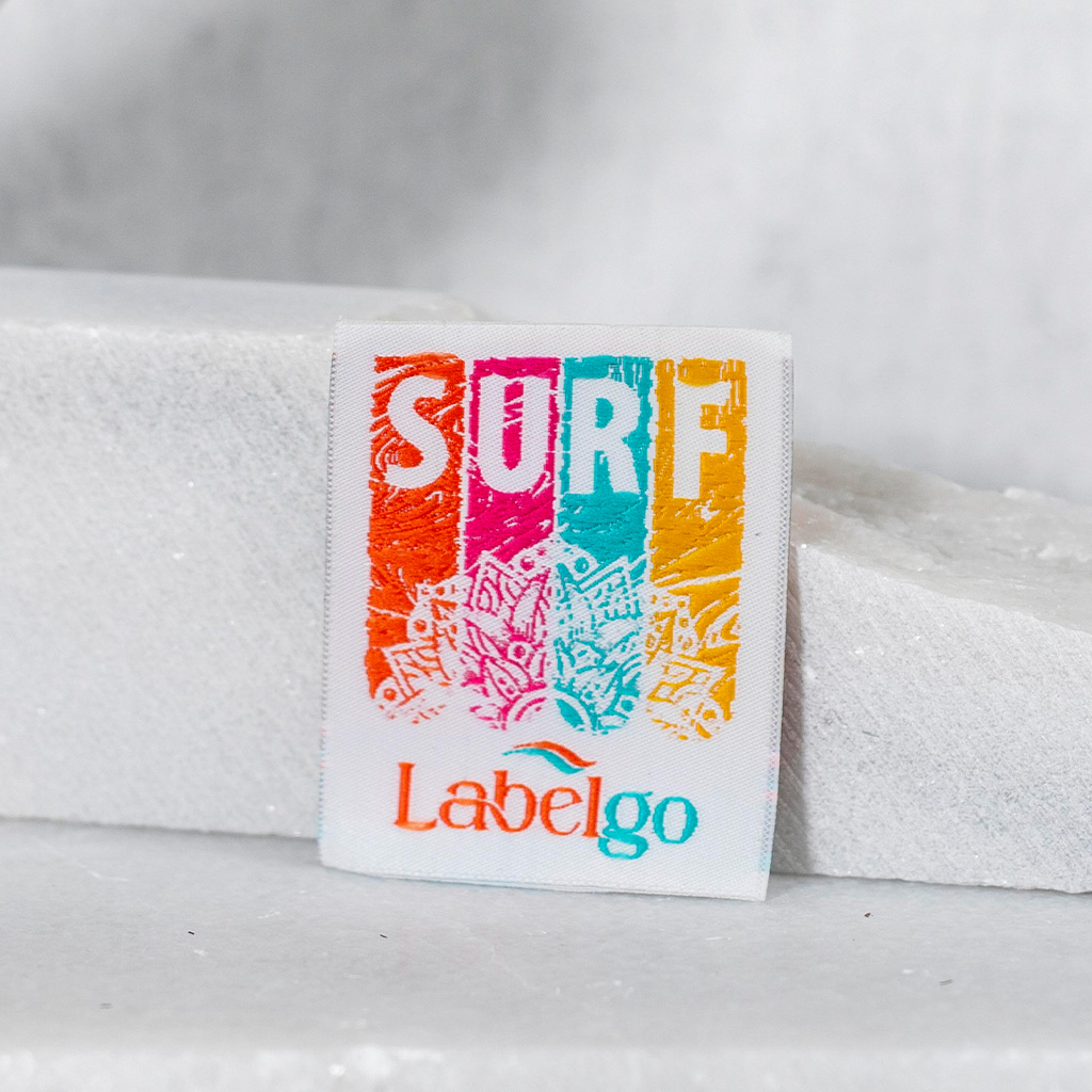 Etiqueta tejida cuadrada con la palabra SURF y diseño colorido de playa.