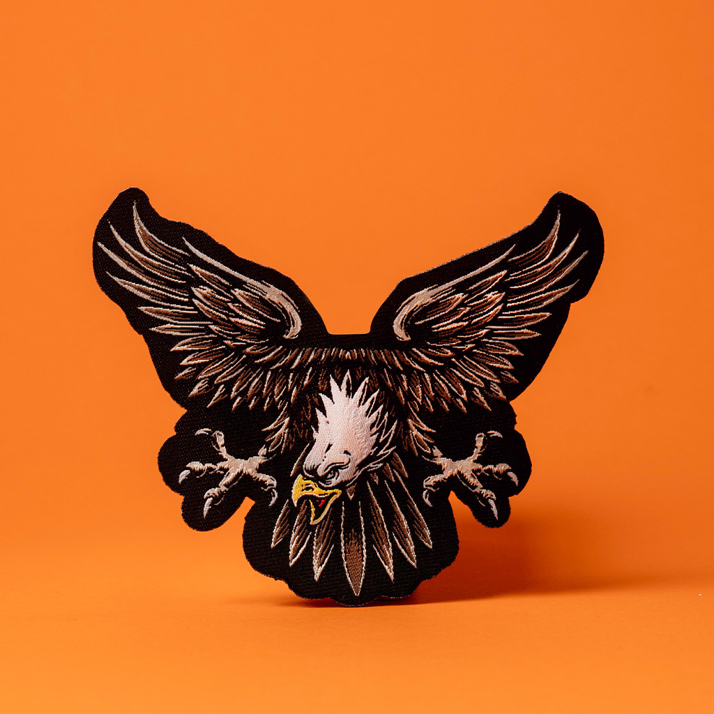 Parche de águila negra bordada con alas abiertas sobre fondo naranja.